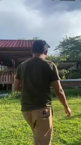 A man wearing a green t-shirt and tan pants walks on a grassy lawn toward a black motorcycle. A young child in a white dress is playing near the motorcycle. In the background, there is a building on a hill and several small wooden structures scattered around the lawn. The scene is viewed from under a corrugated metal roof. The sky is blue with some white clouds.