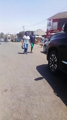 The video shows a street scene in what appears to be a market area.  Two women cross the street in the foreground, followed by a man carrying a green bucket. There are people walking and standing near stalls and shops, with a green and yellow auto rickshaw parked on the side of the road.  The weather appears sunny and clear. Buildings are visible along the street, with signs and flags displayed.
