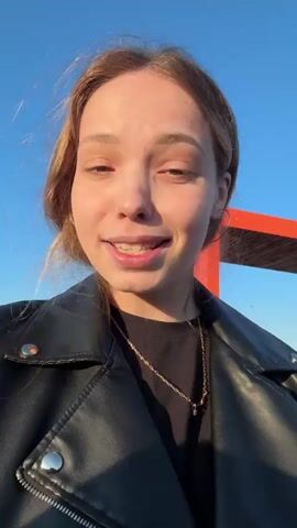 A fair-skinned woman in a black leather jacket is standing outside on a sunny day with a clear blue sky behind her. She has her hair pulled back, wears gold hoop earrings, and a gold chain necklace. An orange and white awning structure is visible in the background. She looks directly at the camera and speaks. She then pulls open the door of a large vehicle, possibly a bus.