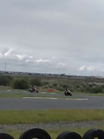 Three motorcycles are on a paved track, driving toward the camera in a left turn. The motorcycles are different colors. One appears to be red, one appears to be black, and another is a mix of black and white. Each motorcycle has a rider in full protective gear. The track is surrounded by green grass and sparse bushes. In the background, there is a cityscape under a cloudy sky.