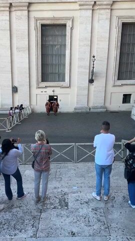 Four people stand on stone steps behind a white fence, facing a courtyard. The people are likely tourists, and are recording the scene with their phones and cameras. In the courtyard, three men in colorful red and yellow striped uniforms with tall hats and spears are performing a changing of the guard ceremony. Another guard in a similar uniform is standing at attention. The courtyard is paved with dark bricks, and surrounded by tall white buildings with large windows.