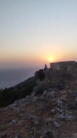 The video shows a sunset over the ocean. A person is sitting on a cliff edge, looking out at the water. There is a small, weathered stone building behind them. The sky is a soft gradient of orange and pink, and the sun is low on the horizon. The foreground of the video is a rocky, uneven terrain with some sparse vegetation. The overall mood of the video is peaceful and contemplative.