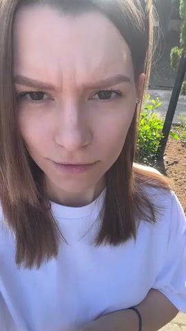 A woman is seen looking at the camera with a slight smile. She is wearing a white shirt and has brown hair. She then places sunglasses on top of her head. The scene takes place outdoors on a sunny day. Trees and foliage are visible in the background. The ground appears to be dirt or mulch.