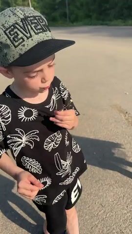 A young boy stands on a paved surface, possibly a road or driveway. He is wearing a baseball cap, a black t-shirt with white tropical designs, and black shorts. He carefully examines a small yellow flower he holds in his hands. The background includes greenery and a blurry shadow of another person. The lighting suggests it's daytime.