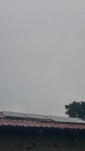 The video displays a gray, overcast sky. A portion of a building with a red roof and windows is visible on the right side of the frame, along with the silhouette of a tree. The building's wall appears to be made of rough stone.