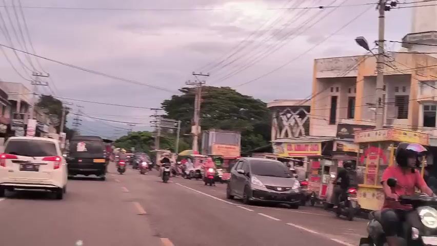The video shows a street scene with various vehicles, including cars and motorcycles, moving along the road. Several food carts are parked alongside the road, and pedestrians are visible. There is a building with a white minaret in the background, and power lines stretch across the sky. The overall atmosphere is that of a bustling urban environment.
