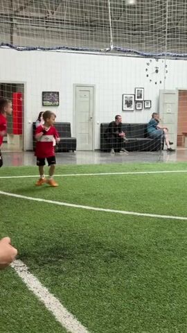 A young boy in a red soccer uniform with the number 10 on his back kicks a soccer ball on an indoor artificial turf field. He is facing away from the camera and towards a soccer goal. Spectators are sitting on couches behind a netting that separates them from the field. The room has white walls, a high ceiling with exposed beams, and several framed pictures on the wall. The boy's left foot is in motion as he kicks the ball, which is in front of him.