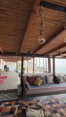 The video pans across a covered patio area with a thatched roof and exposed wooden beams. Two large Edison-style lightbulbs hang from ropes attached to the ceiling. A long, built-in bench with colorful cushions is on the right side of the frame, overlooking the ocean. In the foreground, a mosaic tiled table is visible, with a bag placed on it. The background shows a hallway with white walls and doors, leading to an indoor area. The overall ambiance is relaxed and coastal.