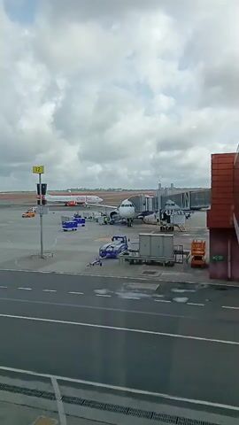 From an indoor vantage point behind a window, the video captures a view of an airport apron on a cloudy day. Two passenger airplanes are visible; the one on the right is parked at a gate with a jet bridge connected to the aircraft. Ground crew personnel are present around this plane, working on what appears to be loading or unloading operations, with ground support equipment such as baggage carts and other service vehicles nearby. The second plane, further away on the left, seems to be parked on the tarmac. A signpost with the number "12" is also visible. The overall scene suggests typical airport activity.