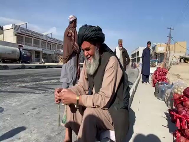 Here's a concise description of the video:

An elderly man with a long white beard, wearing a black turban, a tan shirt, and a dark vest, is sitting on a curb next to a street. He is holding a string of beads in his hands. Behind him, on the sidewalk, there are bags of red onions.  In the background, there are other men walking and buildings lining the street.  A truck is parked across the road. The day is sunny with a clear blue sky.