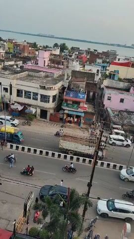 The video provides an aerial view of a busy street scene in what appears to be an Indian city. The street is filled with various vehicles including cars, motorcycles, auto-rickshaws, and a bus. People are seen riding on these vehicles and walking on the sidewalks. Buildings line both sides of the street, with shops and businesses visible at ground level. In the background, a body of water, possibly a river or lake, can be seen, with more buildings extending beyond it. The sky is overcast, suggesting it is either early morning or late afternoon. The overall impression is of a vibrant, urban environment in motion.