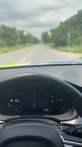 The video captures a first-person view from the driver's seat of a car, traveling on a rural asphalt road, likely in Troitsa, Bulgaria. The road stretches ahead, appearing mostly straight with a slight distant curve, flanked on both sides by dense green trees under a bright, cloudy sky.

The primary focus is the car's interior, showcasing the black multi-functional steering wheel and the digital instrument cluster. The steering wheel prominently features the Volvo logo in the center and various controls, including an "OK" button on the left spoke and cruise control/speed limiter buttons (marked "SET+", "INT", "OFF") on the right.

The digital dashboard provides key driving information. The car's speed is displayed, escalating from 64 km/h to 73 km/h, and then to 80 km/h. The gear is indicated as "D" (Drive), and the drive mode is clearly marked as "HYBRID." A navigation arrow, presumably from a Google Maps integration ("Google" is visible), guides the direction. Other details on the cluster include an outside temperature of 27 degrees Celsius, battery and fuel level indicators, and various mileage and range figures. A small portion of the car's blue exterior (likely the hood) is visible at the base of the windshield. A few distant vehicles can be seen on the road ahead. The scene depicts a daytime drive through a verdant, potentially rural or forested area.