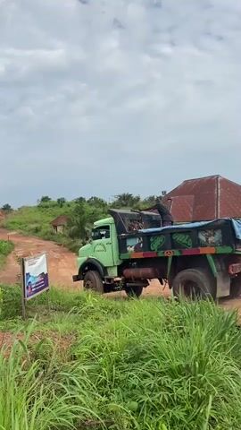 A green dump truck with a blue tarp covering its load moves slowly on a dirt road. A person wearing a dark shirt stands in the bed of the truck, reaching into the load. The truck is surrounded by tall green grass and foliage. In the background, there is a red-roofed building and rolling green hills under a cloudy sky. A sign with an image of a body of water and text is visible on the left side of the road. The scene appears to be in a rural or developing area.
