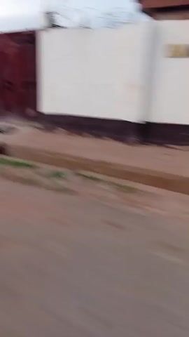 The video appears to be shot from a moving vehicle on a dirt road. The camera pans right revealing a large pile of garbage. A person in dark clothing is seen walking on top of the garbage pile. To the right of the garbage pile is a concrete drainage ditch filled with what appears to be stagnant water and some plants. Beyond the garbage pile is a stone wall, some buildings, and a water tower.