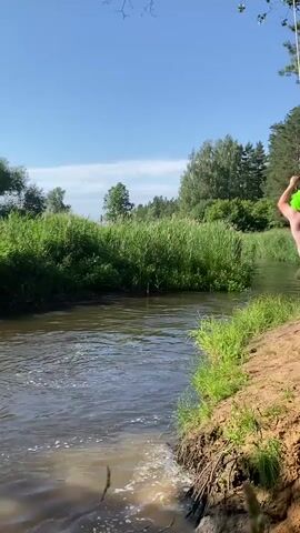 A man wearing a bright green wig and dark shorts is captured mid-air as he swings from a rope over a wide, murky river on a sunny day. The rope is suspended from an unseen point above and to the right of the frame. He is holding onto the rope with both hands, his body arched backward. He is in the process of jumping into the river. Tall, green reeds and grass line the banks of the river, with trees visible in the background under a clear blue sky. The man plunges into the water, creating a large splash and a circular ripple effect.