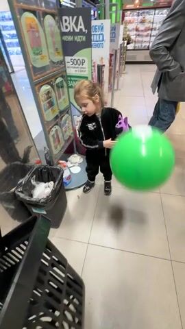 A young child with long brown hair, tied into a ponytail, is walking through a brightly lit store. They are wearing a black tracksuit and holding a purple stuffed toy. The child is looking down and to their left, seemingly engrossed with the toy.

In the foreground, a black shopping basket is visible, its contents blurred due to motion. Behind the child, other shoppers can be seen. One person is holding a large green balloon. The floor is tiled with a light-colored pattern.

The store appears to be a supermarket or a similar retail environment, with shelves of products visible in the background. A display stand with promotional information, including text in Russian, is positioned to the left of the child. The text mentions a discount of "500 рублей" (500 rubles).

The overall impression is a mundane scene of everyday life, likely during daylight hours given the indoor lighting. There are no obvious indications of weather conditions. The activity is simply shopping or walking through the store. The child's focused expression and the presence of a toy suggest a normal, perhaps slightly distracted, moment for a young shopper.