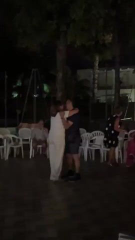 A couple dances closely in the foreground as others sit and watch. The scene is outdoors at night, with trees and some structures visible in the background. The couple is illuminated by a pinkish light, while the rest of the scene is dark.