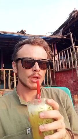 A man with a mustache and sunglasses is sitting in a green plastic chair on a beach. He is drinking a yellow-green beverage with ice and mint from a glass with a red straw. The background shows a building with a red brick base and a wooden porch, as well as a beach with chairs, tables, umbrellas, and palm trees. The sun is setting over the ocean. There are tables and chairs visible further down on the beach. A street light is in the foreground.