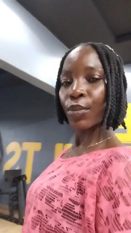 A woman with braided hair, wearing a pink patterned top, stands in front of a mirror in what appears to be a gym. She is looking at the camera and her reflection is visible in the mirror. The reflection shows her from behind, as well as a partial view of the gym equipment behind her. The woman is posing for the camera, with one hand raised to her neck. The lighting is dim and the background is slightly blurred.