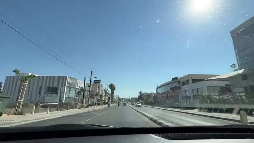 The video shows a first-person view from a car driving down a street on a sunny day. The street is lined with shops and buildings on both sides. There are cars and a scooter on the road. Traffic lights are visible. Palm trees are scattered along the street. The sky is clear blue with a bright sun creating lens flare in the camera. The street appears to be in a commercial or urban area.