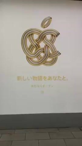 The video shows a promotional poster for Apple in Japan. The poster features the Apple logo rendered in an ornate, interwoven gold and silver design. Beneath the logo are Japanese characters, which translate to "A new story with you. Opening soon." A QR code is printed in the bottom center. The poster is illuminated and appears to be mounted on a wall, with the floor visible below. The camera zooms in slightly on the logo.