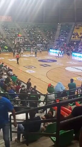 The video shows a basketball game taking place inside an arena filled with spectators.  The court has a light-colored wooden surface with markings and logos. Basketball players are visible on the court, moving around and engaging in game play.  Spectators are seated in bleacher-style seating, many wearing team colors. Some spectators are moving through the aisles.  The arena appears to be well-lit with bright overhead lights. The video seems to be shot from an elevated seating area.  Banners and advertisements are visible in the background.
