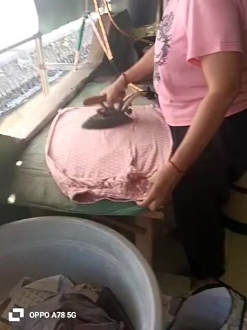 A person wearing a pink shirt and black pants is ironing a pink garment with white polka dots on an ironing board. The garment appears to be a pair of underwear. The person is using a dark-colored iron. The ironing board is covered in a green fabric. In the foreground, there is a large gray basin filled with clothes. The background shows an outdoor environment with some vegetation and a structure that appears to be a roof. The lighting is natural.