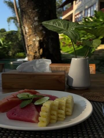 The video shows a breakfast table set outdoors. A plate of cut watermelon and pineapple sits next to a woven place mat, fork, and a coaster. On the coaster is a blue cup of black coffee on a saucer with packets of sugar. A glass of water with a straw is also on the table, along with a wooden tissue box and a gray vase with green leaves. A tree trunk stands behind the table, and a building can be seen in the background amidst green foliage. The table has a woven texture.
