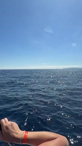 The video shows a view of the ocean from what seems to be a boat or a ship. The water is dark blue with sparkling reflections of the sun. In the distance, there is a land mass on the horizon. The sky is clear blue with a few faint clouds. In the foreground, the arm and thigh of a person are visible, with an orange wristband.