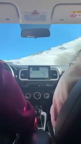 The video shows an interior car view through the front seats. The dashboard features a navigation screen displaying a map and route guidance. The car is driving on a road adjacent to a snowy slope, visible through the windshield under a clear blue sky. A person wearing a magenta-colored sweater is driving, and a passenger is to the right wearing a light-colored hoodie. The focus is on the driving scene and the car's interior.