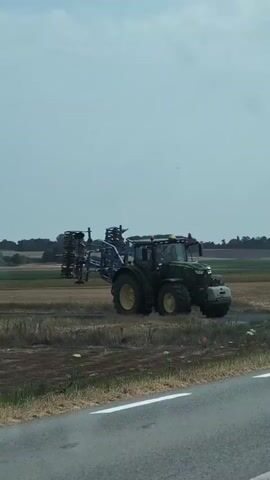 A green tractor with a blue implement attached to its rear is driving on a dirt road next to a paved road. The tractor is turning left at an intersection. A car is driving down the paved road in front of the tractor. The environment appears to be rural with fields and trees. The sky is cloudy.