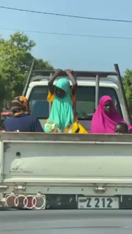 The video shows a white pickup truck with several passengers in the truck bed. The camera is positioned behind the truck, from inside another vehicle. The license plate of the truck is visible, reading "Z 212 MA". The passengers in the truck bed appear to be women wearing colorful hijabs. One woman in a yellow dress and teal hijab is standing and holding onto the truck's frame. The truck is driving on a paved road with buildings and trees in the background, under a clear blue sky. A black bird is visible in the sky. The video is taken from the perspective of someone driving behind the truck.