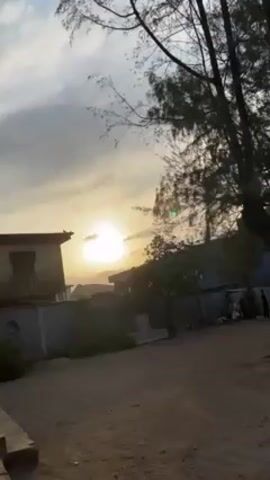 The video shows a view of the sun setting behind a row of buildings and trees. The sky is cloudy, with a mix of light and dark clouds. The sun is a bright orb, partially obscured by the clouds. Some branches of a tree are visible in the right side of the frame. In the lower part of the frame, there's a patch of ground with some bushes and a fence. People are standing in the distance, next to the fence. The overall scene has a warm, golden light, suggesting either sunset or sunrise.