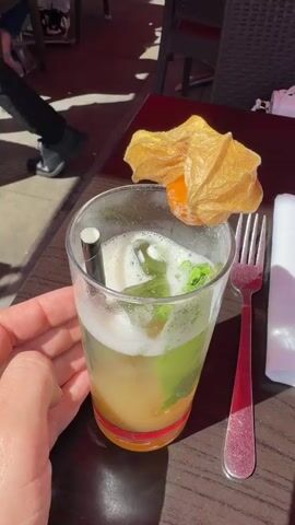 The video showcases a dining scene.  Initially, a hand holds a glass containing a cocktail with a straw, foam, and a golden fruit garnish.  Beside it are a fork and a napkin. The next shot shows a plate of spaghetti carbonara with parsley garnish.  The video then widens to show a table with three plates of pasta dishes, one a spaghetti with a tomato sauce and basil garnish, one a brown sauce pasta dish, and a piece of Extra gum. A mobile phone rests beside the plates on the table.  A person in a white shirt sits at the table. The table and setting suggest an outdoor restaurant environment.
