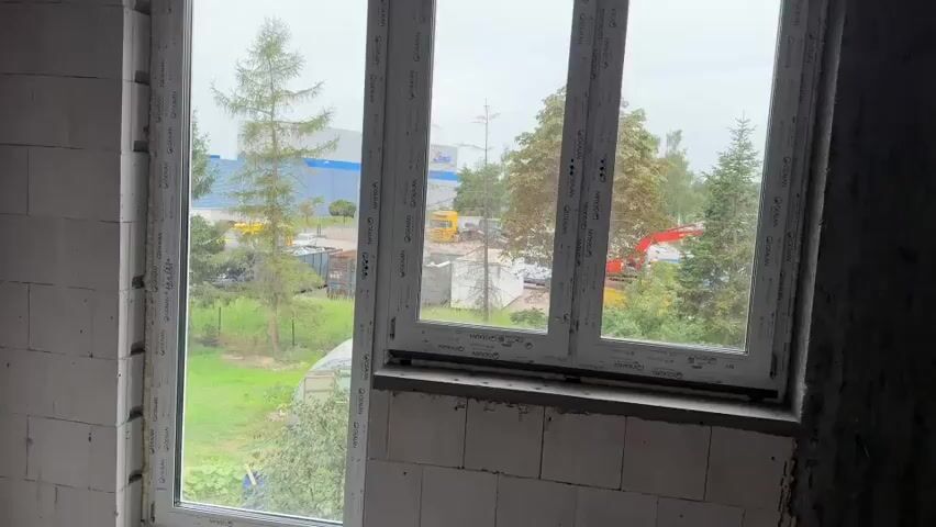The video pans across an interior construction site. The walls are made of white cinder blocks. There are windows with white frames that look out onto a green, outdoor area with trees, a yellow truck, and construction equipment. There are electrical conduits and outlets visible on the walls. The floor is covered with debris.