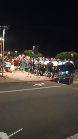 A group of musicians, mostly men, sit in green plastic chairs on a paved area at night. They are playing instruments, including what appear to be brass instruments like tubas and trombones, as well as drums on the left side. A speaker on a tall black stand is positioned in front of the group. There are music stands in front of many of the musicians. A conductor, wearing a dark shirt, stands on the far left, gesturing with his hands. Another person, also in dark clothing, stands on the far right, appearing to direct the musicians. The background is dark, with trees visible behind the musicians. The foreground shows a dark asphalt road with a white pedestrian crossing symbol painted on it. There are some road lights visible in the distance. The scene appears to be an outdoor performance.