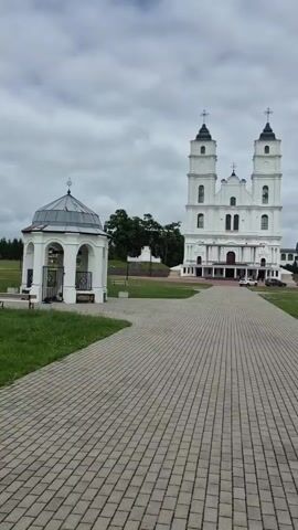 The video captures a walking perspective of a grand white church with two prominent towers, a smaller gazebo-like structure to the left, and a long, paved pathway. The church, likely the Aglona Basilica, stands majestically against a cloudy, overcast sky. The pathway is laid with rectangular pavers and curves towards the right, leading the viewer's eye towards the church and a spacious green lawn. The lawn is dotted with sparse white wildflowers and a few benches.

To the right of the church, another white building with a dark roof is visible, appearing to be part of the church complex or a related institution. Trees are scattered across the landscape, with a denser cluster of trees further in the background. The overall atmosphere is serene and somewhat solemn due to the overcast weather.

The time of day appears to be daytime, but the cloud cover obscures direct sunlight, creating a diffused light. No people are explicitly visible in the frame, suggesting a quiet or off-peak visiting time. The scene is set in a prominent religious or historical site, characterized by its impressive architecture and manicured grounds. The paving stones and the neat lawn suggest a well-maintained tourist or pilgrimage destination. There is no visible text in the video.