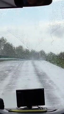 The video, taken from inside a vehicle, shows a road covered in rain and puddles. Water droplets are visible on the windshield. Trees line the sides of the road. In the distance, a truck is driving in the same direction. A small black screen is mounted on the dashboard. A windshield wiper is seen sweeping across the glass. The sky is overcast.