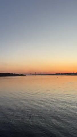 The video shows a body of water with gentle waves under a gradient sky that transitions from a light blue at the top to an orange and yellow hue near the horizon. Silhouetted against the horizon are trees on the left bank, a distant bridge with tall towers, and some buildings or structures on the right bank. The overall scene depicts a calm sunset or sunrise over a wide expanse of water.