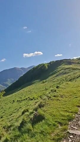 The video showcases a scenic landscape of rolling green hills and distant mountains under a clear blue sky. The camera pans across the terrain, revealing lush meadows and forested slopes. A dirt path winds through the landscape, and the shadows of two people walking along it are visible. A man wearing sunglasses, a grey and black jacket, and a backpack comes into view, holding a can in his hand. He glances to the side as he stands on the path.