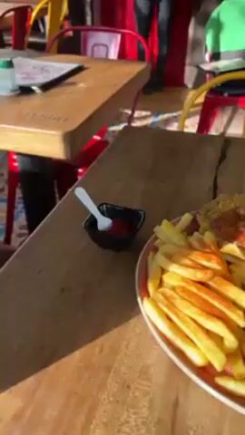 The video shows a restaurant setting, starting with a close-up of a plate containing french fries, a fried plantain with sauce, and a breaded meat dish topped with dark sauce. The camera pans to reveal a table with condiments, a yellow smoothie-type drink, and a foil-wrapped item. The restaurant has wooden tables and colorful chairs. There is writing on the wall, "Asadero".  The last shot shows a person taking pictures of the meal with a pink cell phone. There are people sitting at other tables in the background. The restaurant appears to have both indoor and outdoor seating areas.