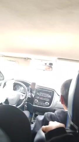 A video shows the interior of a car. The camera is positioned in the back seat, looking forward. The driver is visible in the driver's seat, with hands on the steering wheel. The dashboard of the car is also visible, including the radio and a phone mounted on a stand. Outside the car, trees and a road are visible.