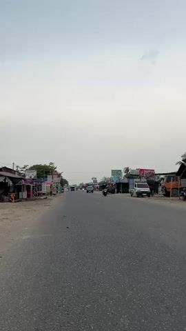 The video shows a view of a two-lane asphalt road with shops lining the sides. The sky is overcast with clouds. Several vehicles, including cars and motorcycles, are traveling on the road. On the left side of the road, there are various small shops with colorful signs. On the right side, there are also shops and some parked vehicles. The overall scene depicts a busy roadside environment.