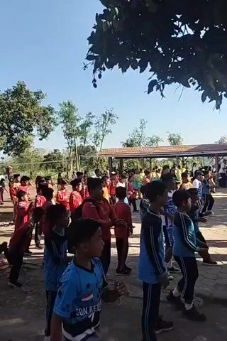 A group of children, mostly boys, in matching blue and black athletic wear, stand in rows outdoors on a paved area. They are performing exercises. Some children have their arms raised, while others appear to be marching or moving their arms. In the background, there is a building with a red roof and a covered walkway. There are also trees with green leaves and a clear blue sky. The children are of various ages, appearing to be elementary school aged. The sun is shining brightly, casting shadows on the ground.
