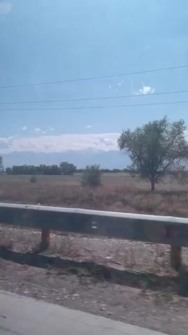 A wide, static shot shows a rural landscape under a clear blue sky with scattered clouds. In the foreground, a metal guardrail runs horizontally across the bottom of the frame, casting a shadow. Beyond the guardrail, a dry, grassy field stretches towards a distant line of trees and snow-capped mountains. A utility pole with wires is visible on the left side of the image. The overall impression is one of a vast, open, and undeveloped area.