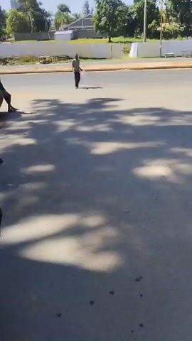 The video shows a street scene in an urban area.  The road is wide with some traffic including motorcycles and a person walking. A large boulder is on the side of the road. One person sits on it, and another stands nearby. Further away, another person walks and several people are running towards the boulder. The day appears sunny.
