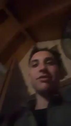 A close-up selfie-style video features a young man singing and gesturing towards a woman lying in a bed in the background. The man has short, dark hair, light skin, and is wearing a dark shirt and red jacket. He appears excited, smiling, and opening his mouth wide as he sings. The woman in the bed has long, dark hair and is partially covered by a white sheet. She is smiling and looking at the man. The room appears dimly lit, with a wooden ceiling and walls. A window can be seen in the background. A red object is visible on a nightstand near the bed. The camera movement is shaky and follows the man's gestures and facial expressions.