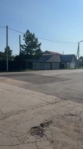 This video shows a rural or suburban street scene. The camera pans from left to right, showing a cracked concrete sidewalk in the foreground. Beyond the sidewalk is an asphalt road with some patches. On the right side of the road, there are several garages or outbuildings with grey siding and a reddish roof. A tall evergreen tree stands in front of the buildings. Further back, there are houses and fences, with more trees and vegetation. The sky is clear and blue, with a bright sun visible in the upper left corner of the last frame. There are no people visible in the video. The overall impression is a quiet, sunny day in a somewhat weathered environment.