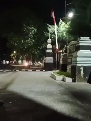 The video shows a nighttime scene at the entrance of a building labeled "DPRD". The building appears to be an official government or legislative institution. A flag, predominantly red and white, is displayed on a flagpole. The entrance is marked by a gate or barrier, and the area is lit by artificial lights, creating shadows on the road. Trees and foliage are visible in the background. The road is paved, and there are white and black markings along the side of the road. In the distance, vehicle headlights can be seen, suggesting traffic. The camera is positioned at a low angle, looking towards the entrance. There are no clear individuals visible in the frame, though a person's legs can be seen in the lower left corner. The overall atmosphere is dark and somewhat stark.