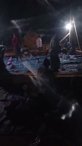 The video shows a group of people gathered outdoors at night. A bright light source, possibly a portable floodlight on a stand, illuminates the scene. The ground is covered with blue tarpaulin, and there are several people seated on the tarpaulin, appearing to be eating or socializing. Some individuals are standing and interacting with others. There are purple plastic bags and food items scattered on the ground. In the background, there seems to be a structure, possibly a building or a temporary shelter, made of wood or similar materials. The overall atmosphere suggests a communal gathering or event taking place after dark.